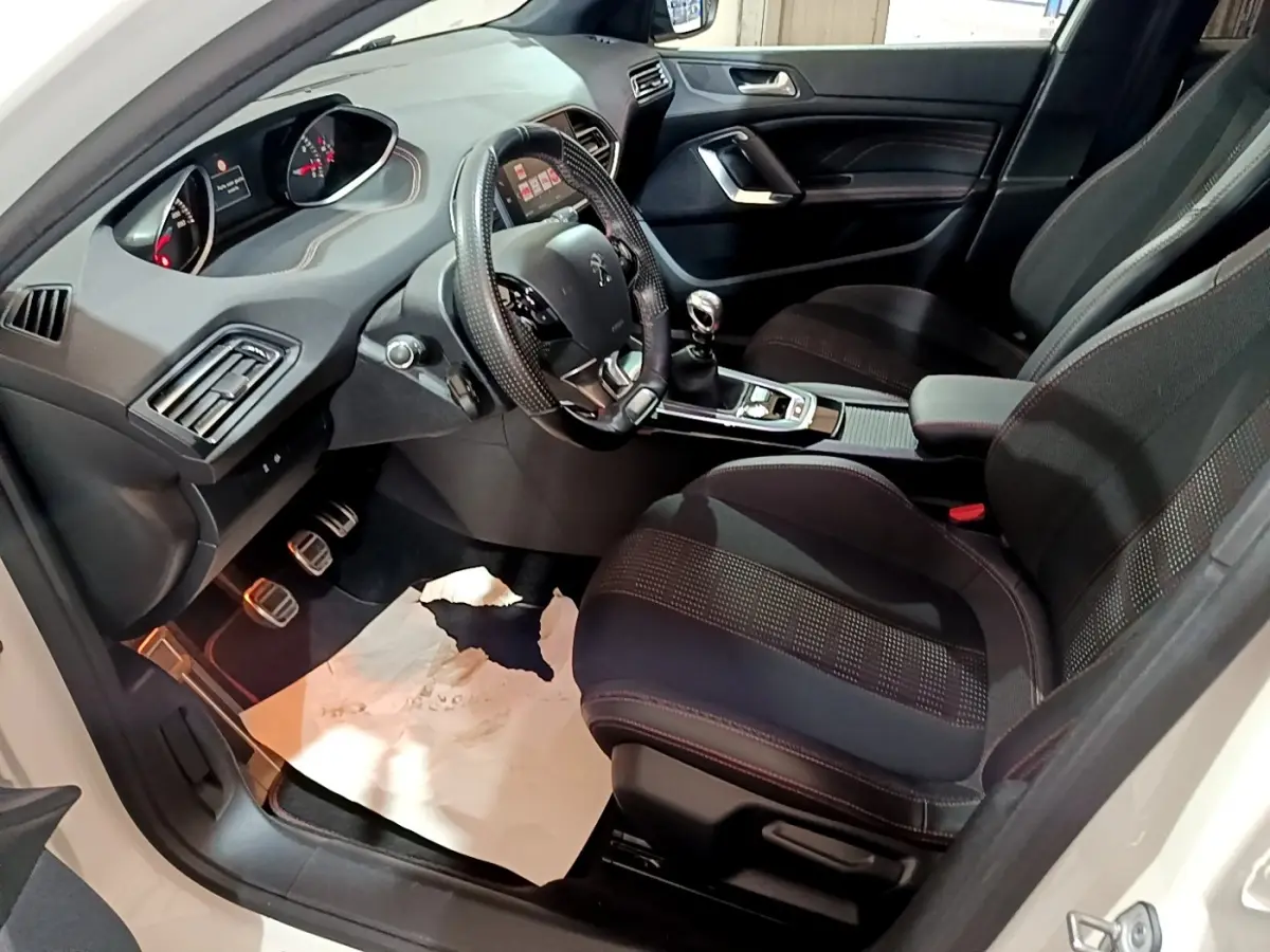 Vue intérieure côté conducteur de la Peugeot 308 GT Line blanche, montrant le volant cuir, la boîte manuelle et les sièges noirs.