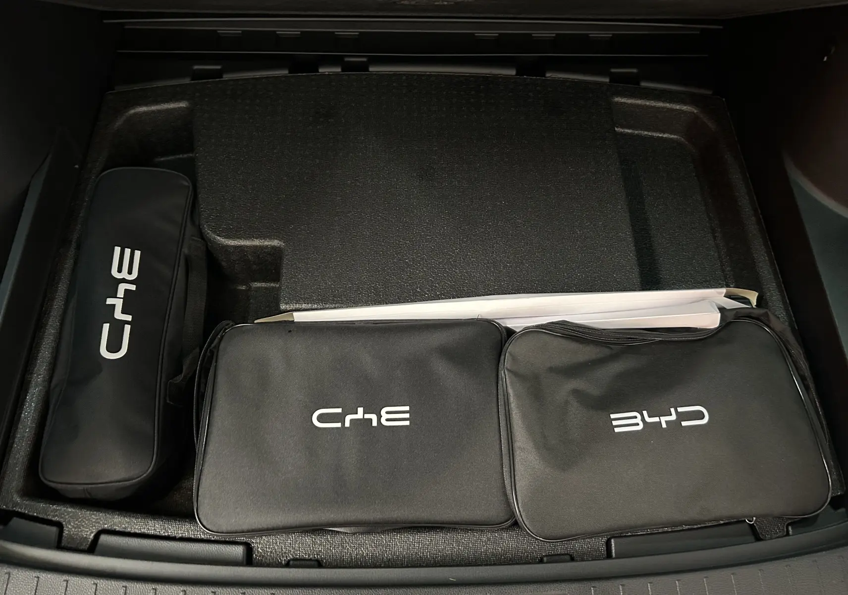 Vue plongeante sur le coffre avec trois trousses noires de marque BYD et ChE, sur fond intérieur noir.