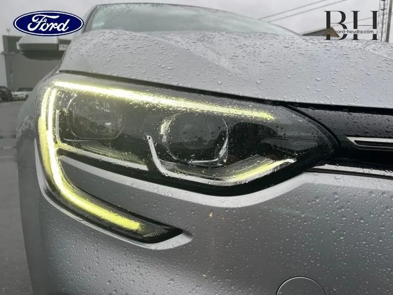 Gros plan sur le phare avant gauche d'une Renault Mégane Break gris platine sous la pluie, avec éclairage LED allumé.