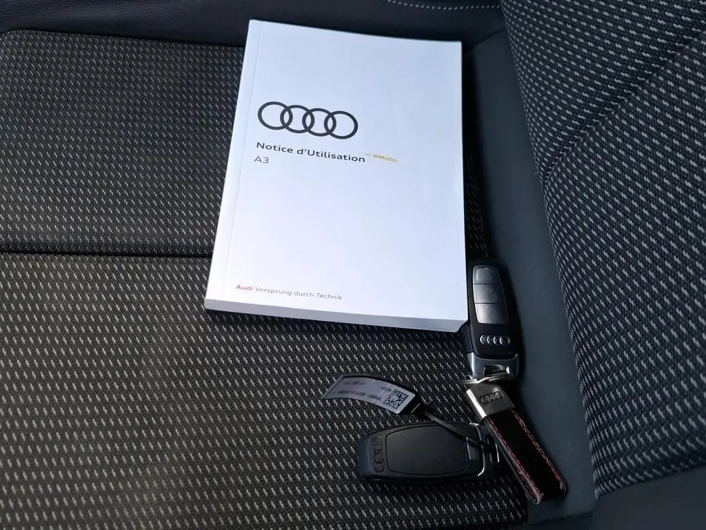 Notice d'utilisation Audi A3 posée sur un siège tissu noir à motifs avec une clé de voiture Audi à côté.