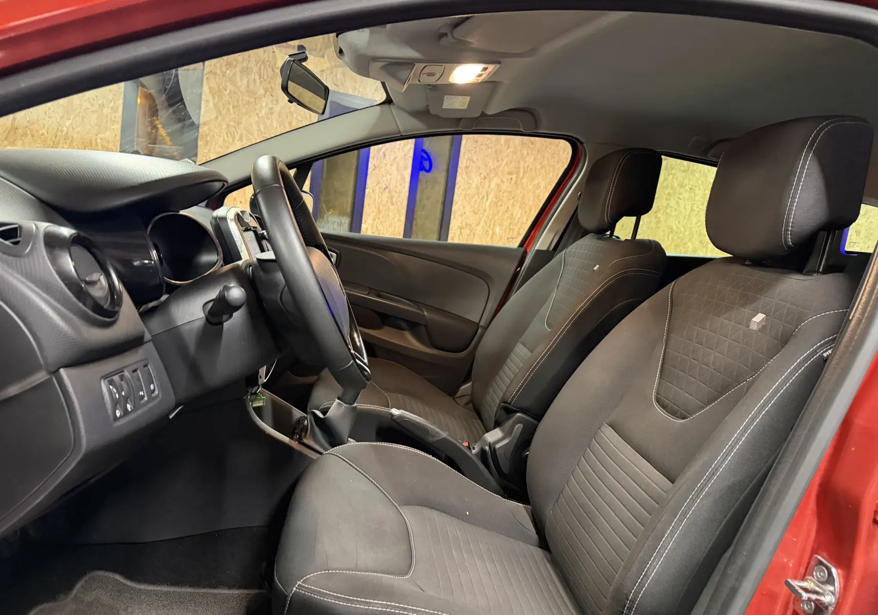 Intérieur avant droit de la Renault Clio Estate IV rouge, montrant les sièges noirs et le tableau de bord avec volant et commandes.