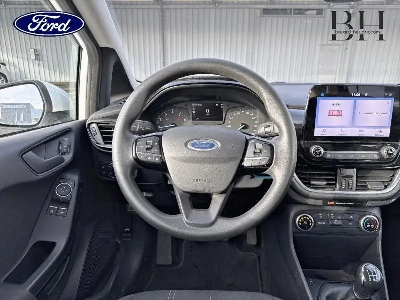 Vue intérieure centrée sur le volant et le tableau de bord de la Ford Fiesta blanc Glacier avec écran tactile et commandes au volant.