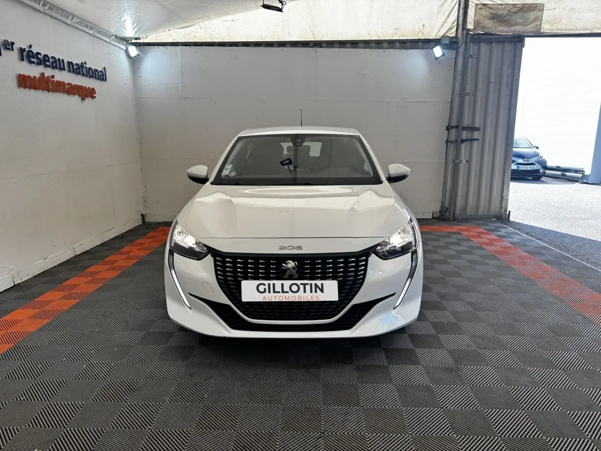 Vue avant d'une Peugeot 208 blanche stationnée en intérieur, avec calandre noire et phares allumés.