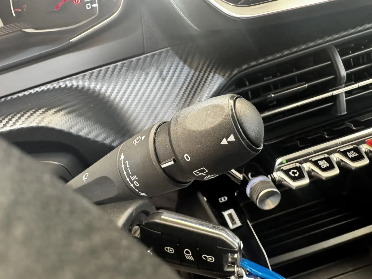 Gros plan sur la commande d’essuie-glace noire et la console centrale avec boutons et finition carbone dans l’habitacle d’une Peugeot 208 blanche.