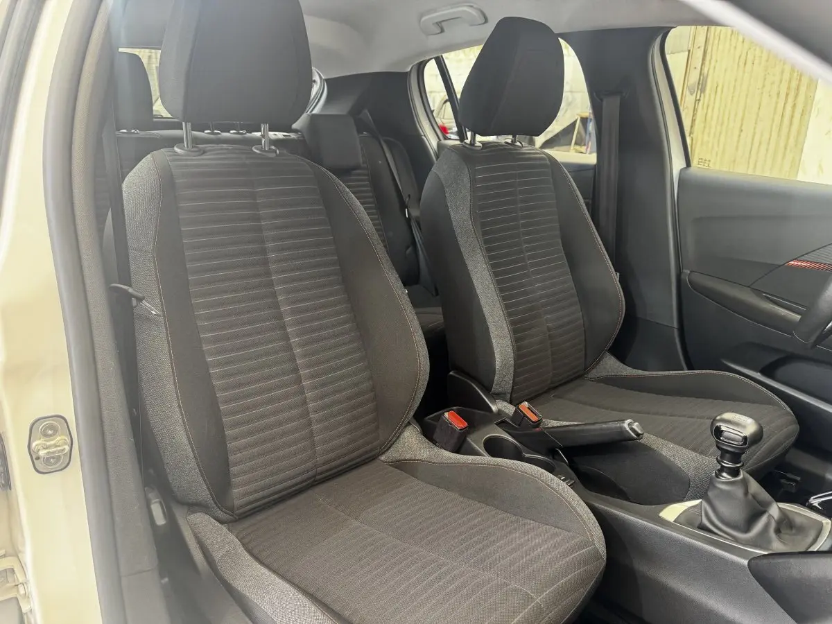 Vue intérieure côté avant droit de la Peugeot 208 blanche, montrant les sièges tissu noirs et la boîte manuelle.