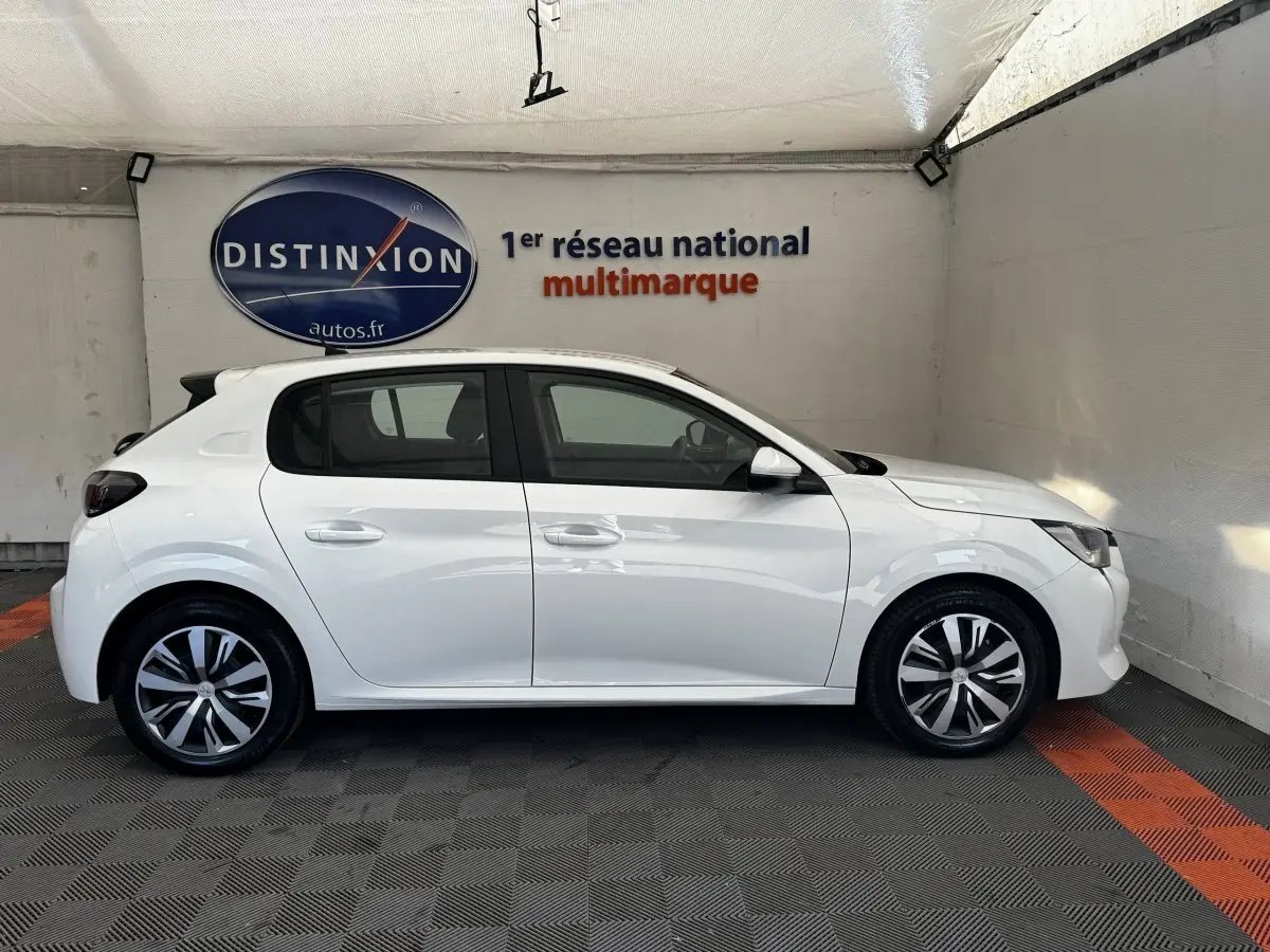 Profil latéral droit d'une Peugeot 208 blanche stationnée en intérieur, avec jantes alliage et vitres teintées.