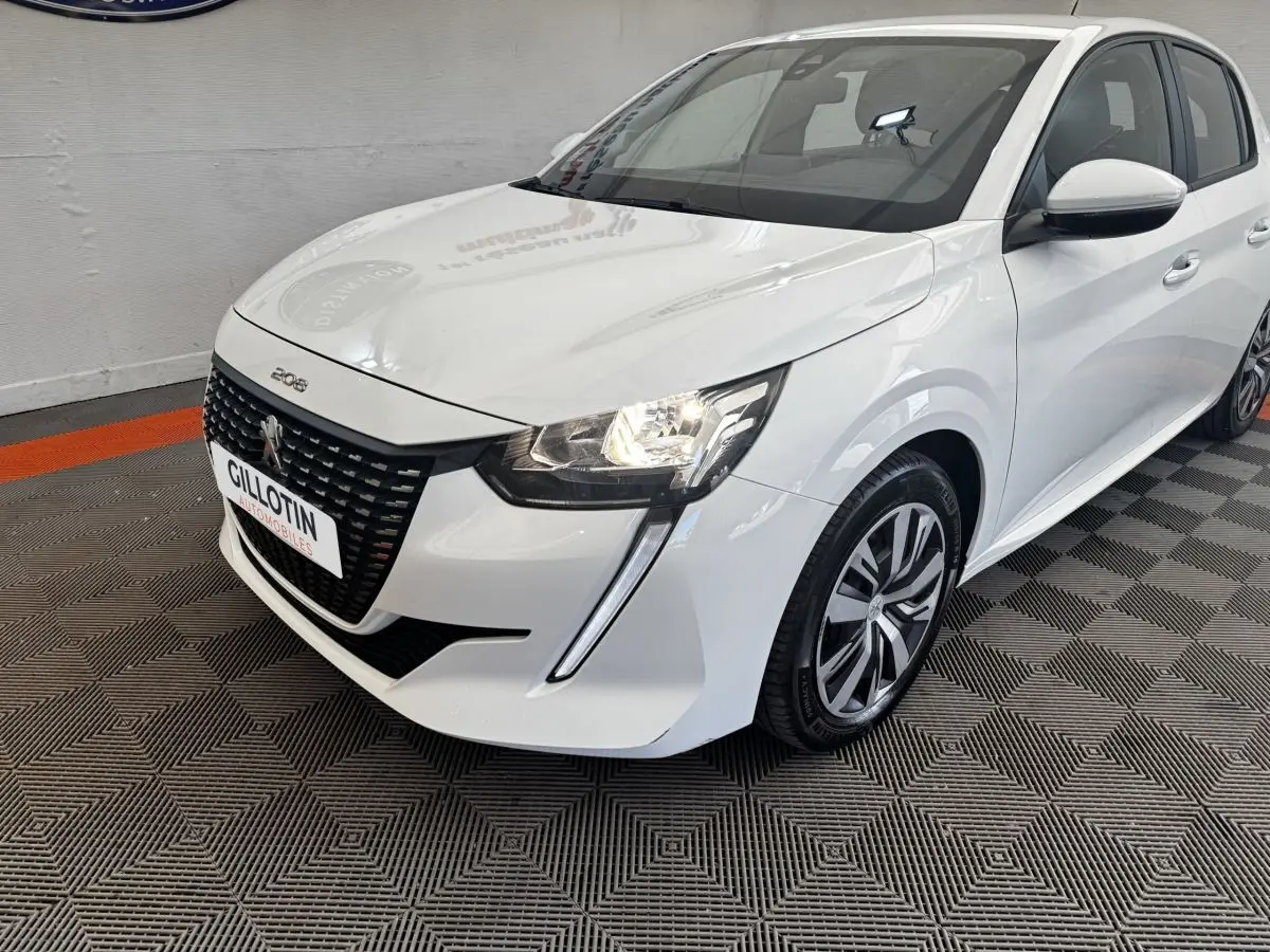Peugeot 208 blanc vue 3/4 avant droit, avec calandre noire distinctive et feux allumés dans un showroom.