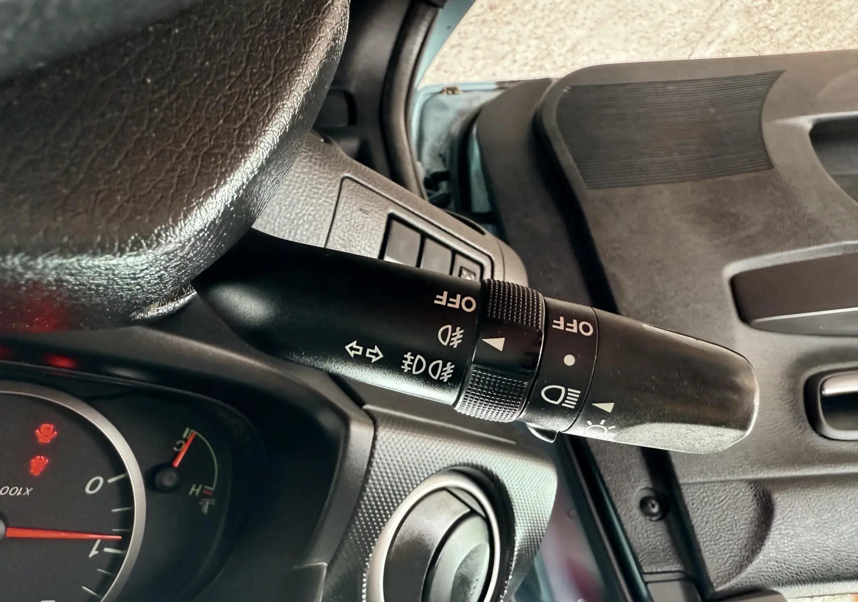 Gros plan sur la manette de clignotants et phares à gauche du volant dans l'habitacle noir d'une Mazda2 gris 2004.