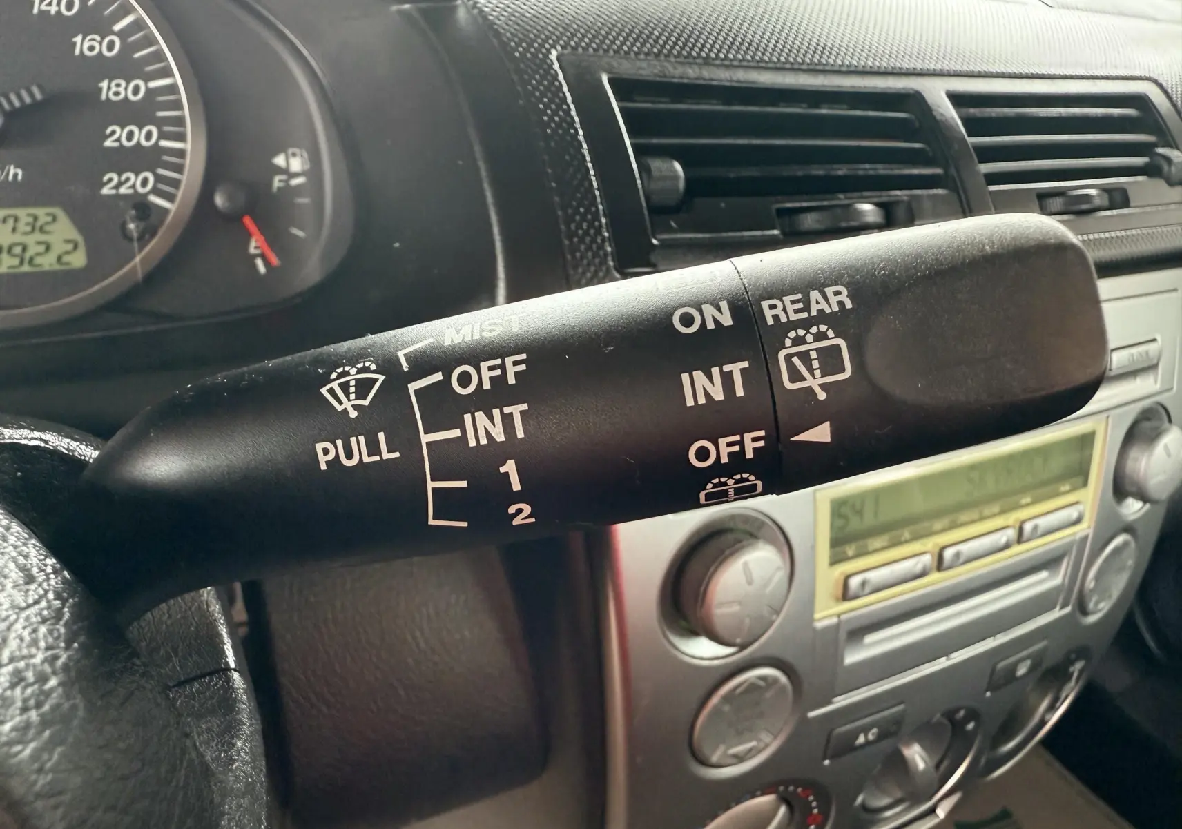 Gros plan sur la manette d'essuie-glace côté gauche du tableau de bord d'une Mazda2 gris 2004, avec commandes visibles.