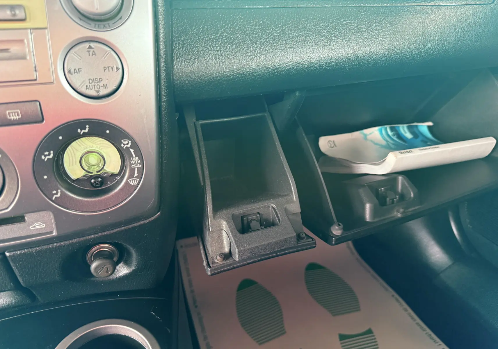 Intérieur de la Mazda2 2004 montrant la console centrale et la boîte à gants ouverte avec manuel visible.