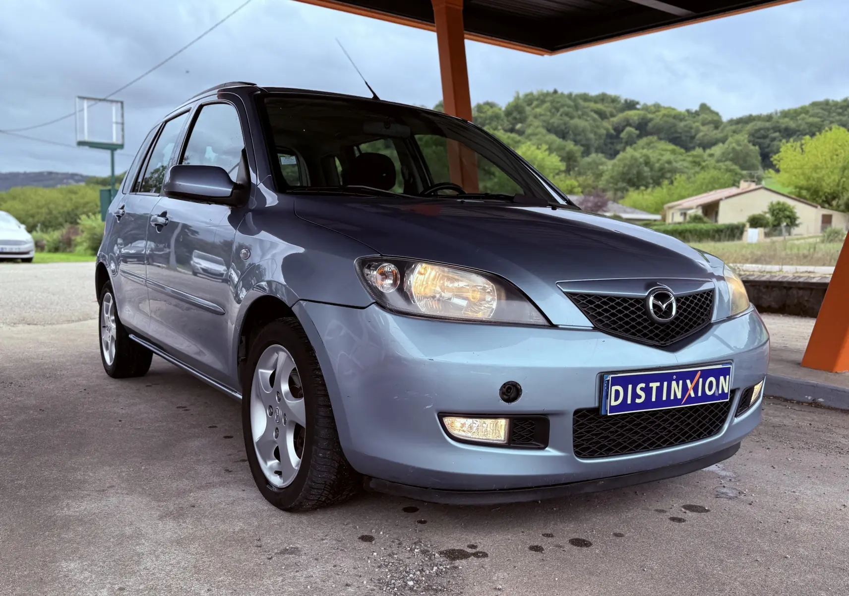 Vue 3/4 avant d'une Mazda2 gris métallisé de 2004 sous abri, avec phares allumés et jantes alliage.