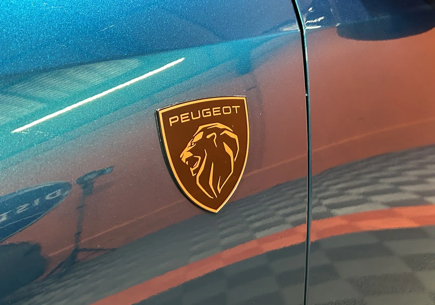 Gros plan sur l'écusson Peugeot doré sur la carrosserie bleu obsession du côté gauche du Peugeot 5008 III Hybrid 145 GT.