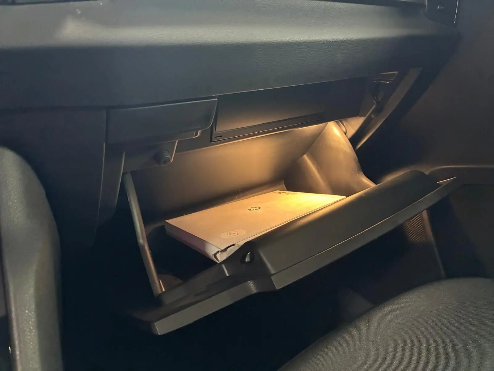 Gros plan intérieur sur la boîte à gants ouverte éclairée du Skoda Karoq gris 2023, avec manuel visible.