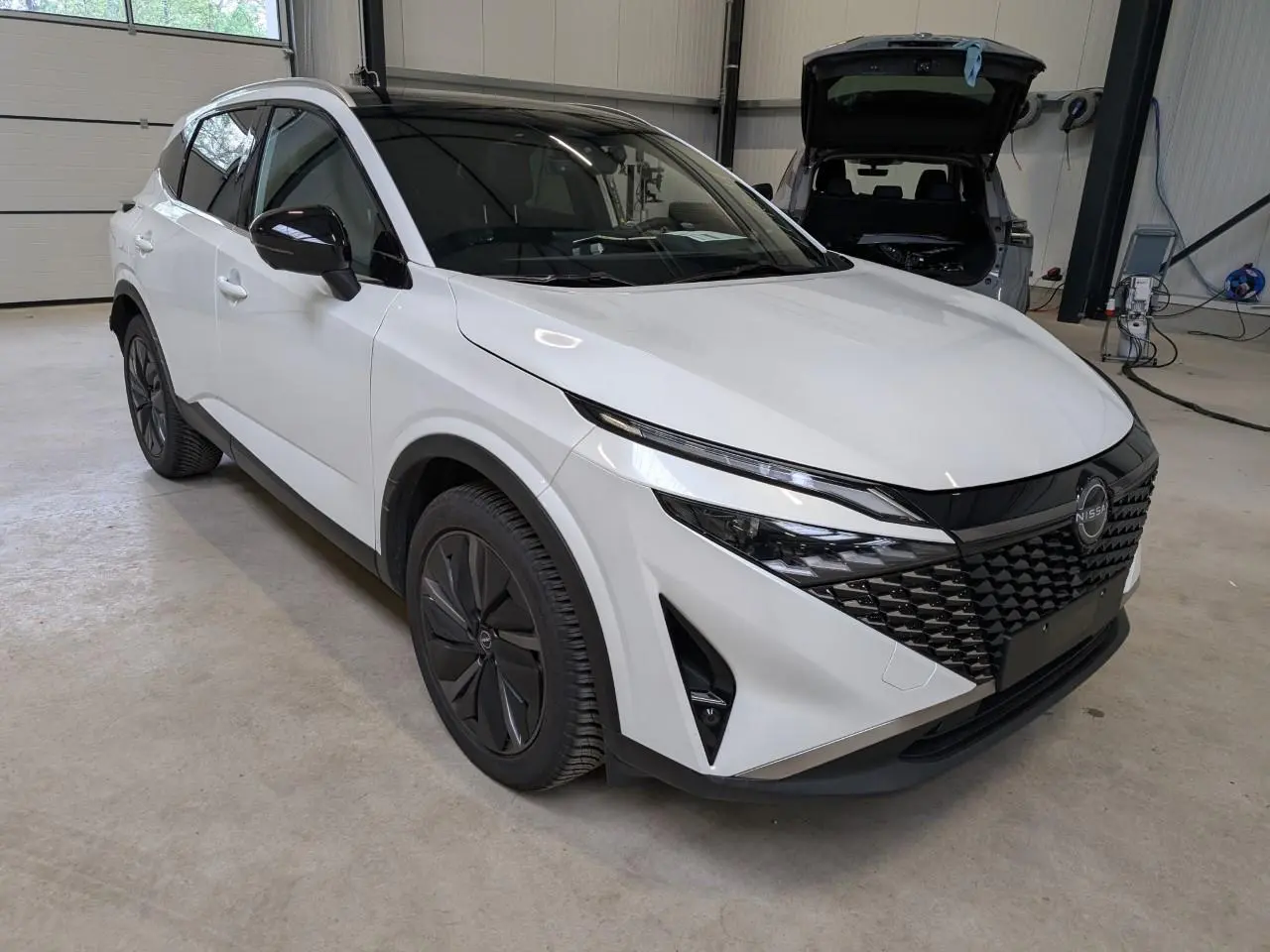 Nissan Qashqai blanc nacré vu en 3/4 avant droit, avec calandre noire et jantes alliage foncé dans un garage.