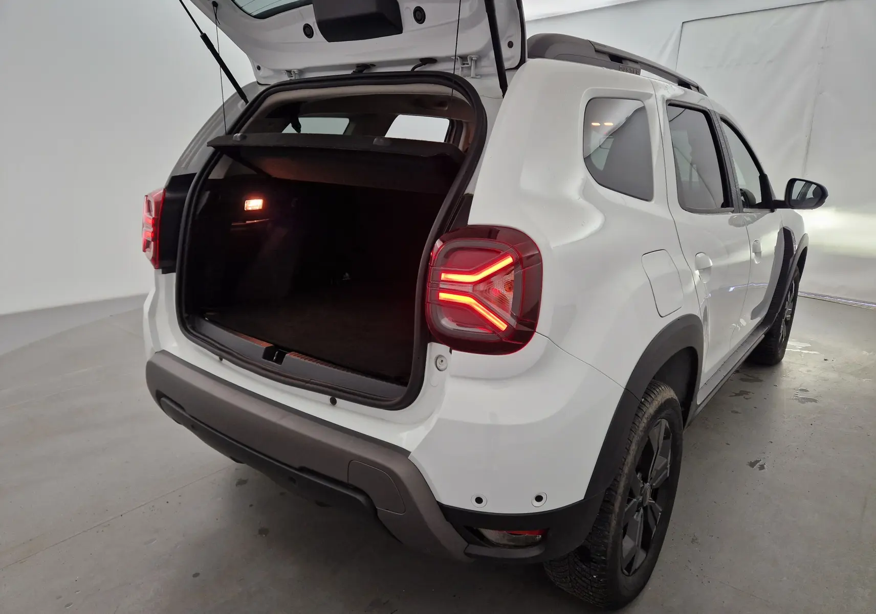 Vue arrière droite d'un Dacia Duster blanc 2024 avec coffre ouvert et feux arrière allumés dans un studio.