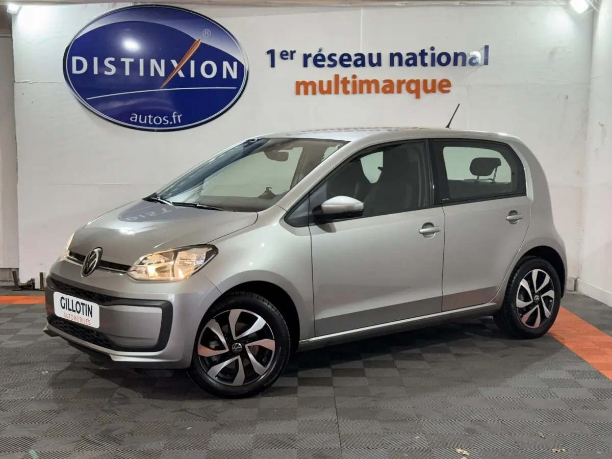 Volkswagen Up gris clair vue de profil côté droit, feux allumés, jantes noires et argentées en intérieur showroom.