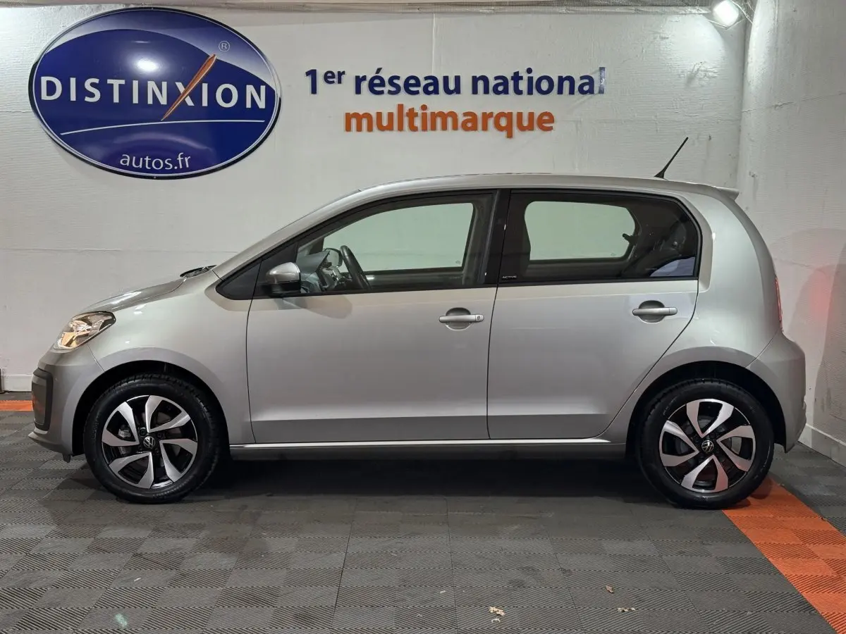 Profil droit d'une Volkswagen Up 1.0 65 Active gris foncé, avec jantes alliage et vitres teintées, en intérieur showroom.