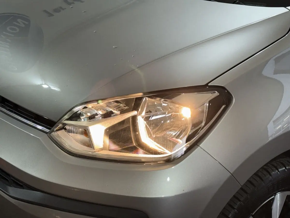 Gros plan sur le phare avant droit allumé d'une Volkswagen Up gris foncé, avec partie du capot et aile visible.