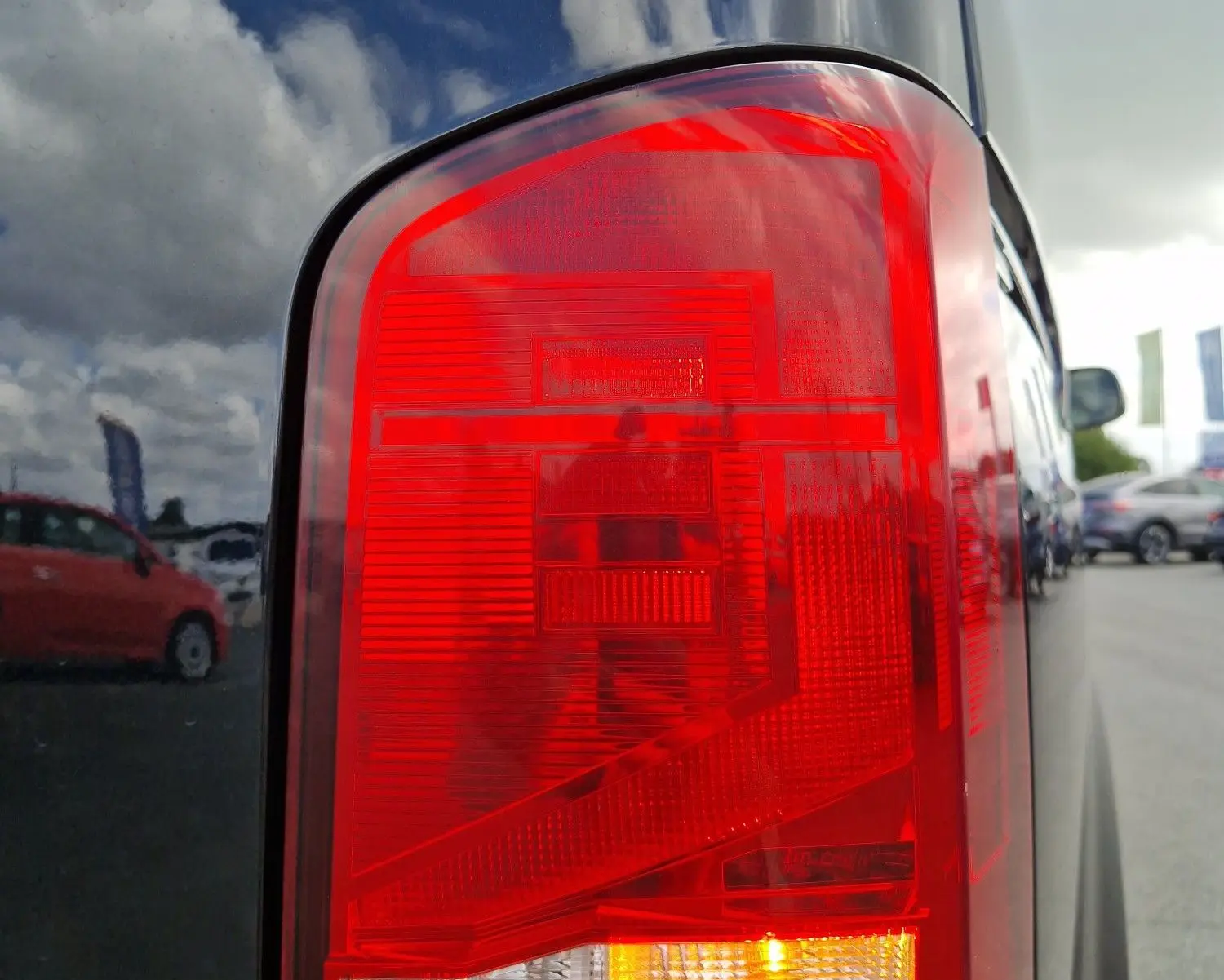 Gros plan sur le feu arrière droit rouge et orange d'un Volkswagen Transporter noir, reflet du ciel et véhicules alentours visible.