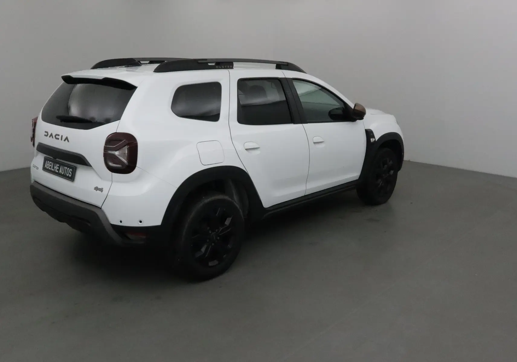 Vue 3/4 arrière droite d'un Dacia Duster blanc 4x4 avec jantes noires et barres de toit dans un studio.