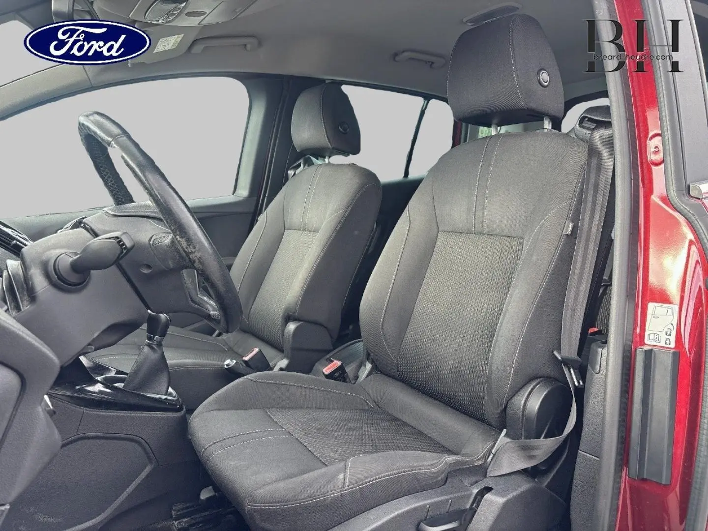 Vue intérieure côté conducteur du Ford B-MAX rouge Premium Candy, montrant les sièges avant en tissu gris et le volant cuir.