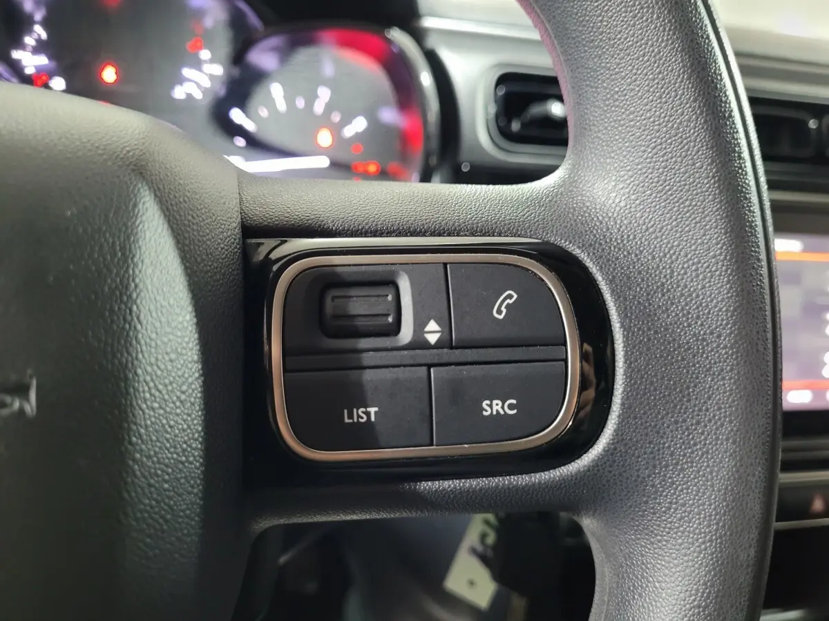 Gros plan sur les commandes gauche du volant noir de la Citroën C3 beige, avec tableau de bord et écran tactile en arrière-plan flou.