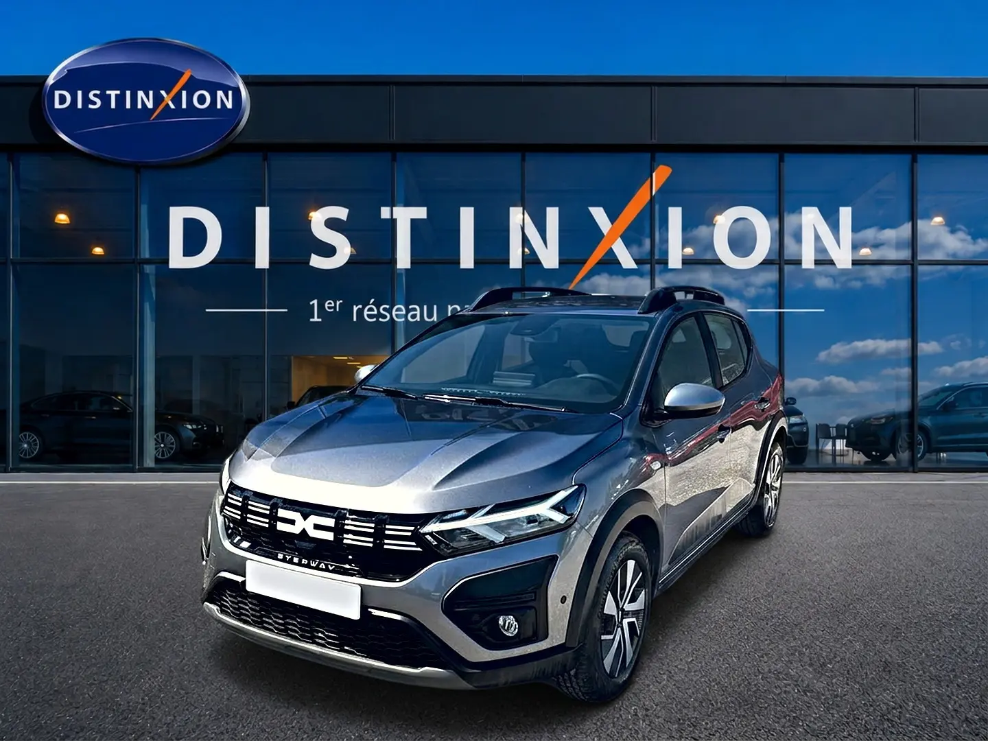 Dacia Sandero Stepway gris schiste vue 3/4 avant droit avec barres de toit devant un bâtiment Distinxion.