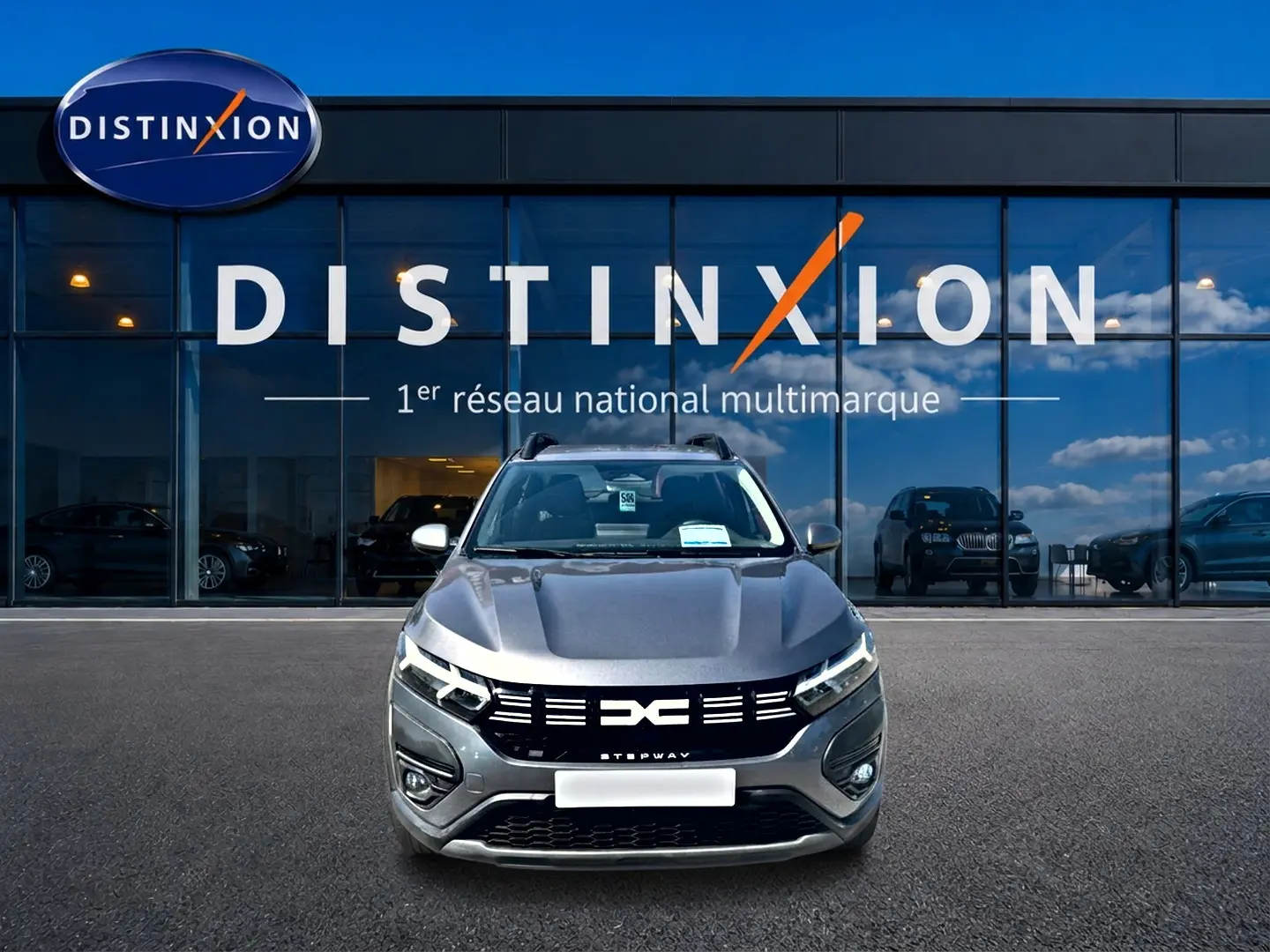 Vue avant d'une Dacia Sandero Stepway gris schiste avec calandre noire et barres de toit devant une vitrine Distinxion.