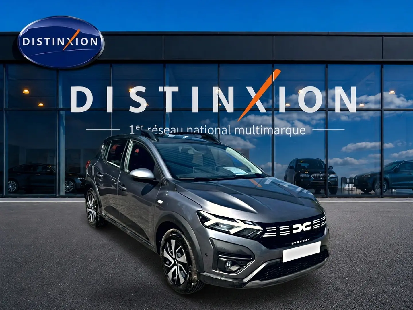 Vue 3/4 avant droit d'une Dacia Sandero Stepway gris schiste avec barres de toit et jantes distinctives devant un showroom Distinxion.