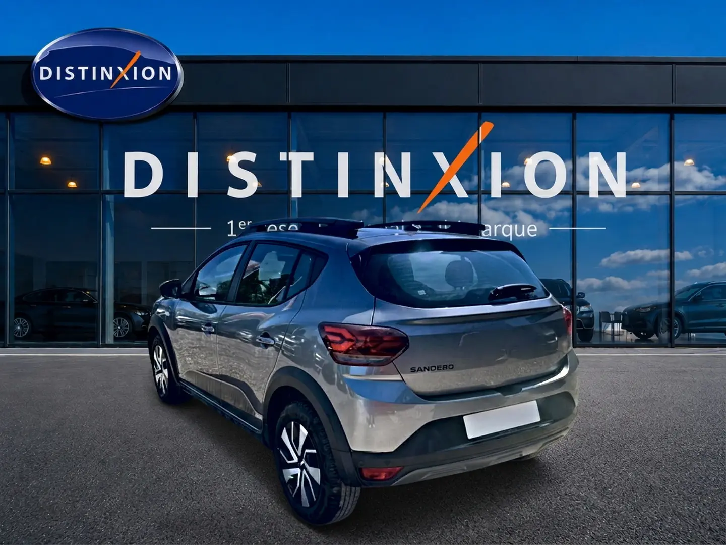 Vue 3/4 arrière droite d'une Dacia Sandero gris schiste avec barres de toit et jantes distinctives, devant un showroom Distinxion.
