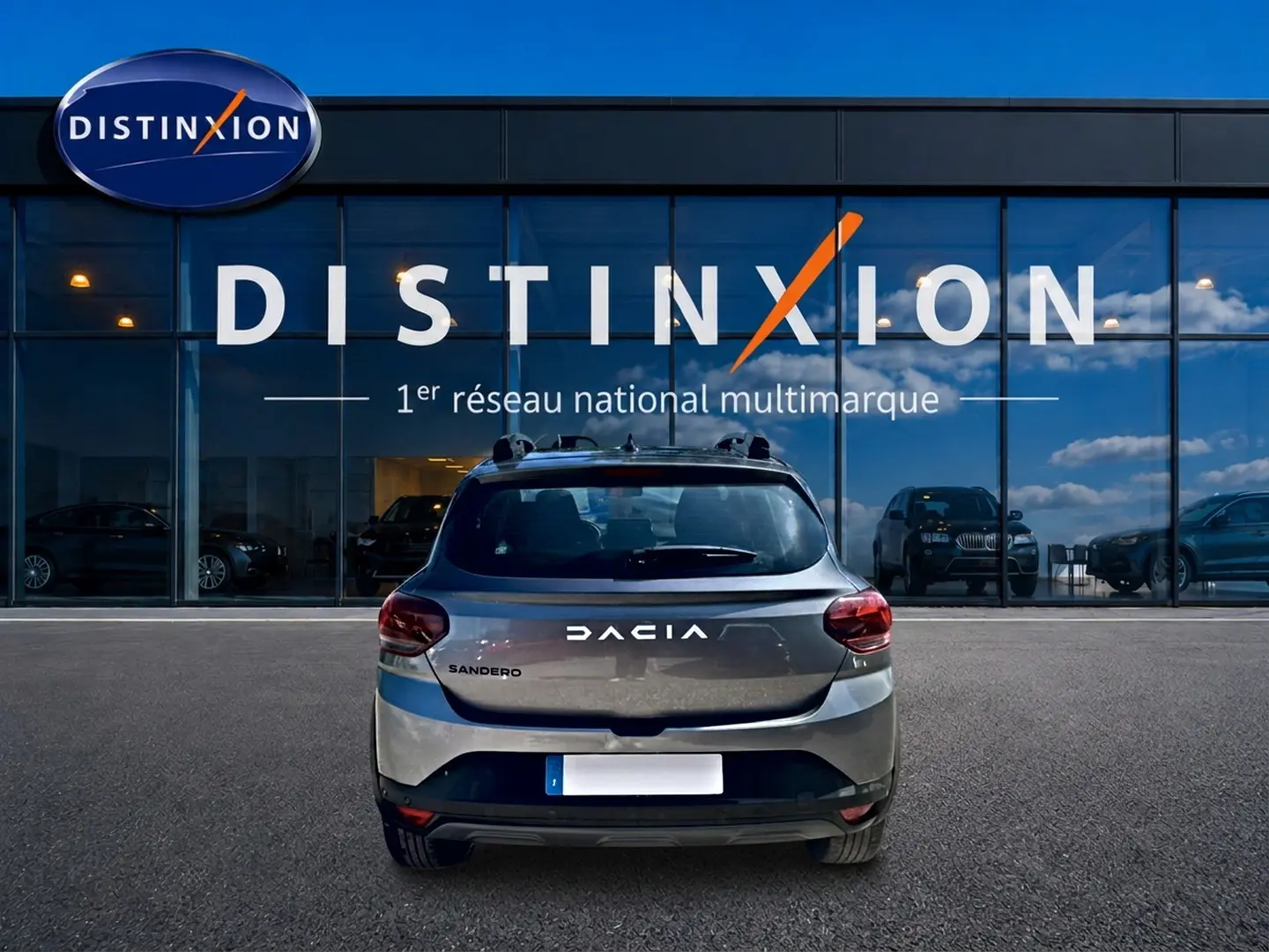 Vue arrière d'une Dacia Sandero gris schiste 2025 avec barres de toit et logo distinctif sur fond de vitrine Distinxion.