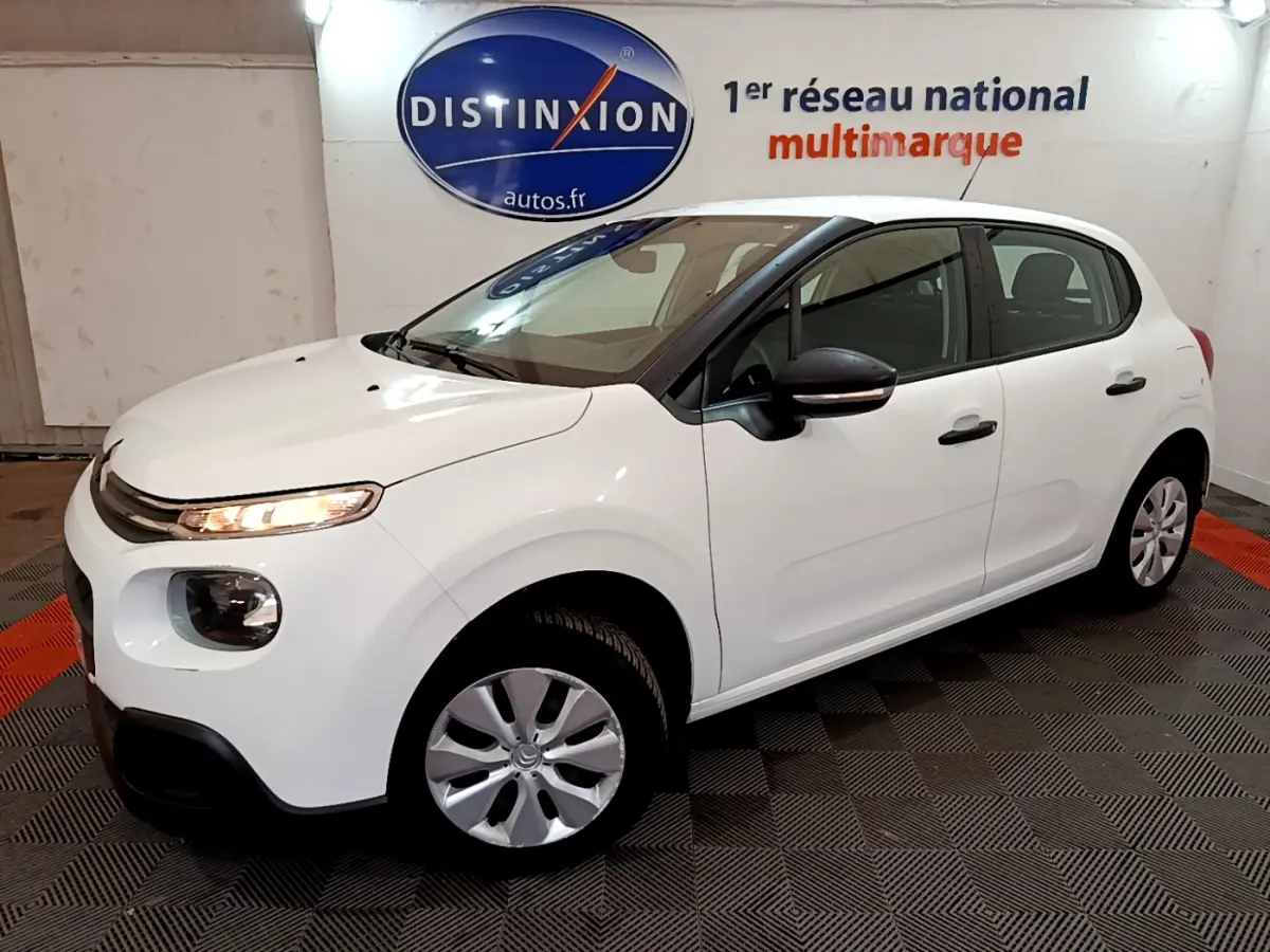 Citroën C3 blanche vue en 3/4 avant droit, avec feux allumés et rétroviseurs noirs dans un showroom.