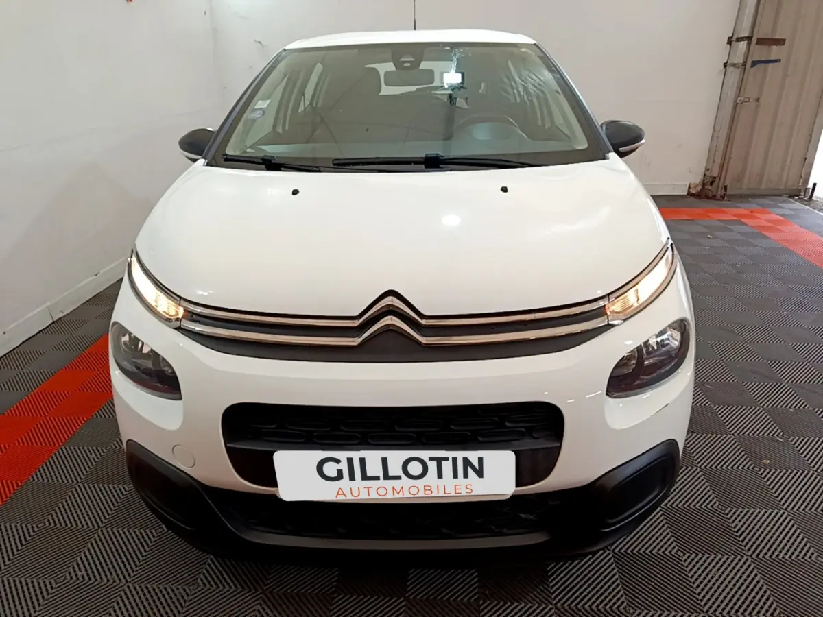 Vue frontale d'une Citroën C3 blanche de 2018 avec phares allumés et calandre distinctive en intérieur showroom.