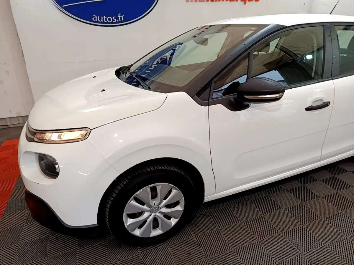 Vue 3/4 avant gauche d'une Citroën C3 blanche avec rétroviseurs noirs et jantes argentées en intérieur showroom.