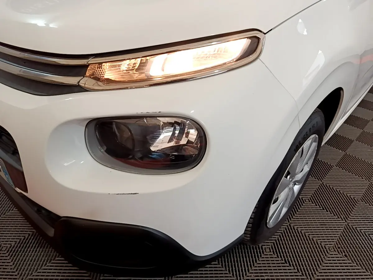 Gros plan sur l'avant droit d'une Citroën C3 blanche de 2018, mettant en valeur ses phares allumés et la roue.