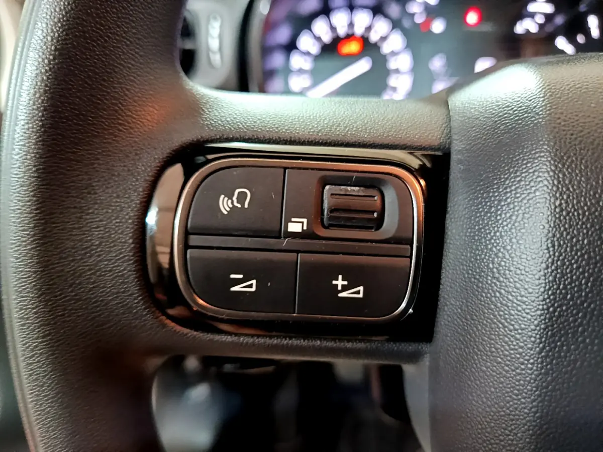 Gros plan sur les commandes multifonctions noires du volant d'une Citroën C3 beige, avec tableau de bord flou en arrière-plan.