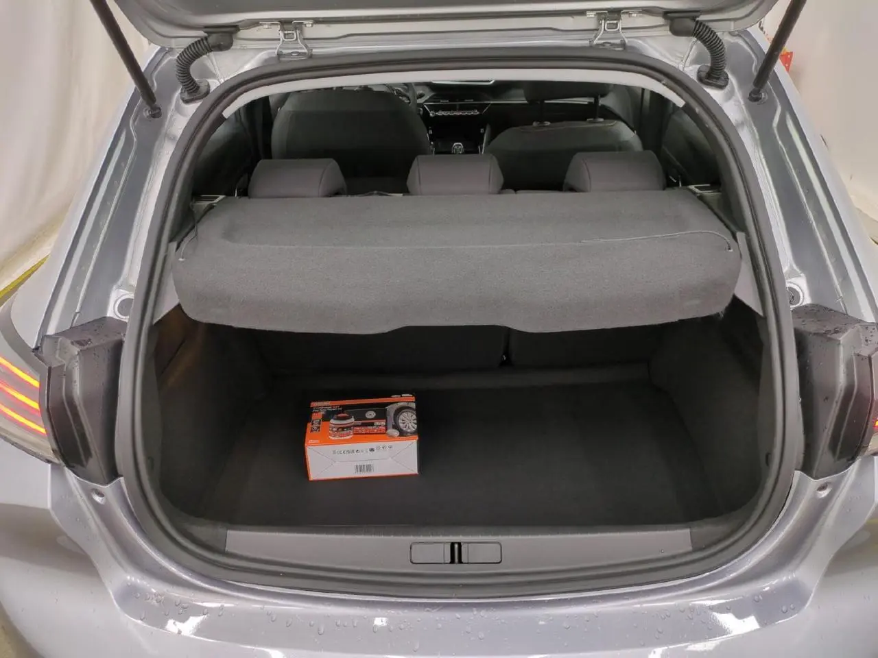 Coffre ouvert d’une Peugeot 208 gris clair, vue arrière montrant un kit de dépannage pneumatique posé au sol.
