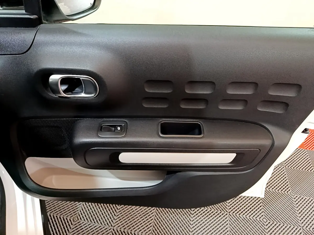 Vue rapprochée de la porte intérieure côté gauche d'une Citroën C3 blanche avec poignée argentée et panneau noir texturé.