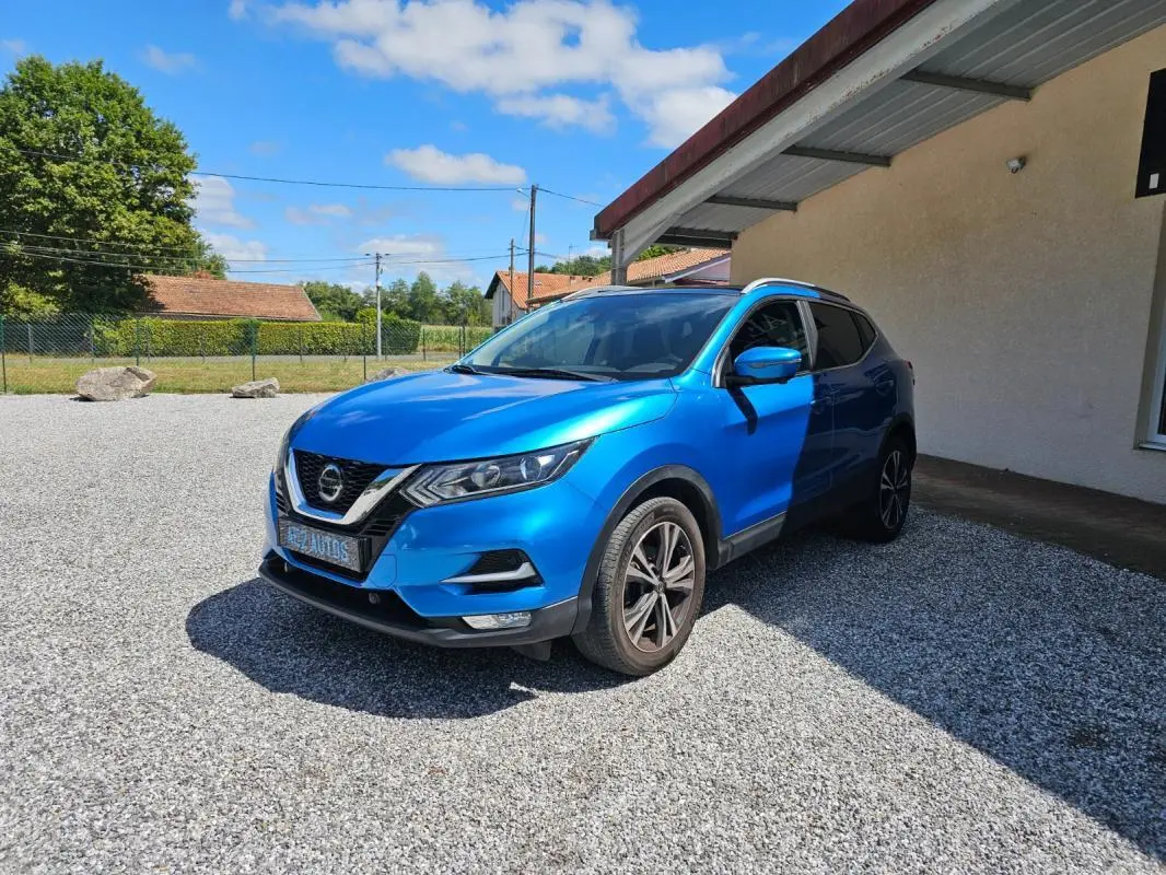 Nissan Qashqai bleu 2019 en 3/4 avant droit, stationné sur gravier devant un bâtiment beige.