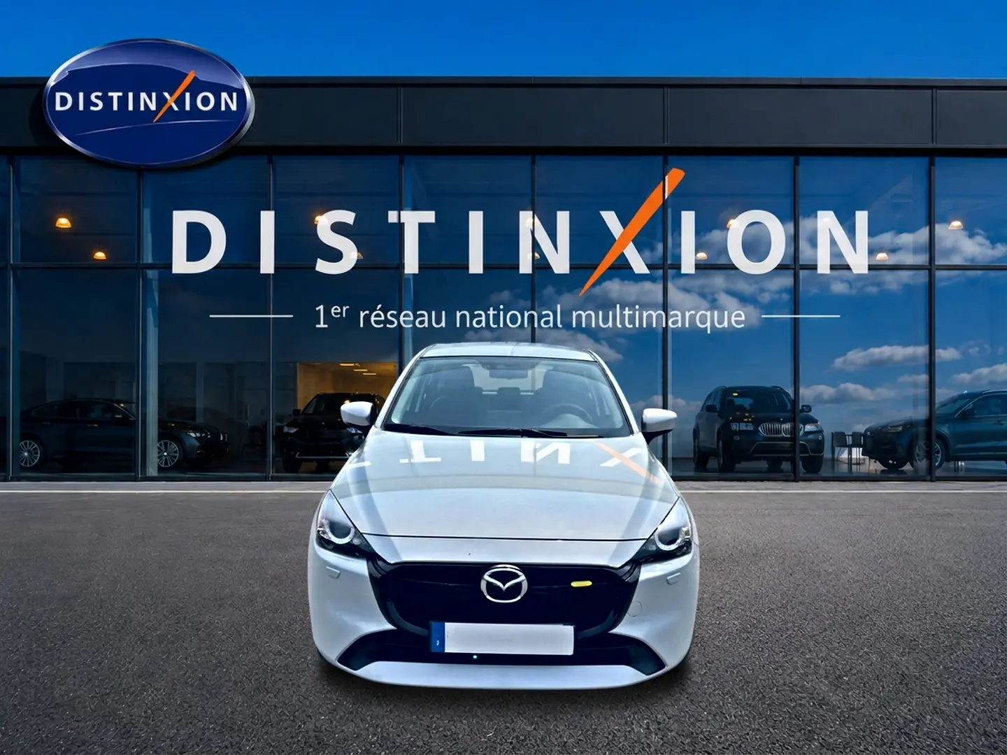 Vue frontale d'une Mazda2 blanche 2024 garée devant une vitrine avec logo Distinxion en reflet sur le capot.
