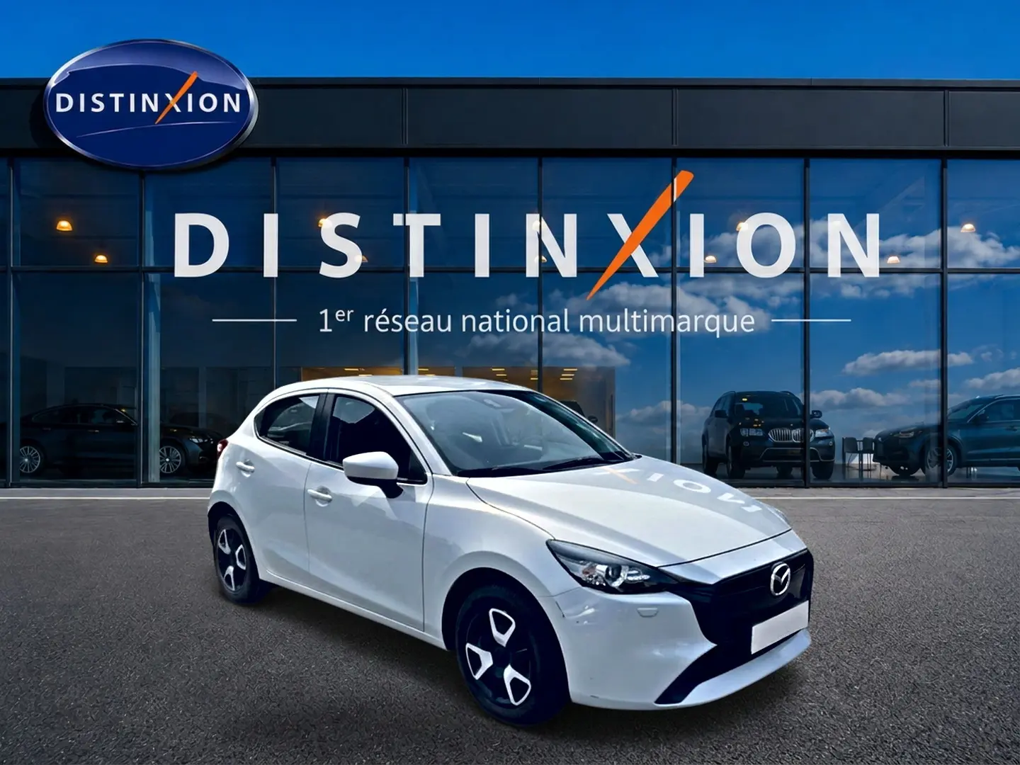 Vue 3/4 avant d'une Mazda2 blanche 2024 avec jantes noires et calandre noire devant un showroom Distinxion.