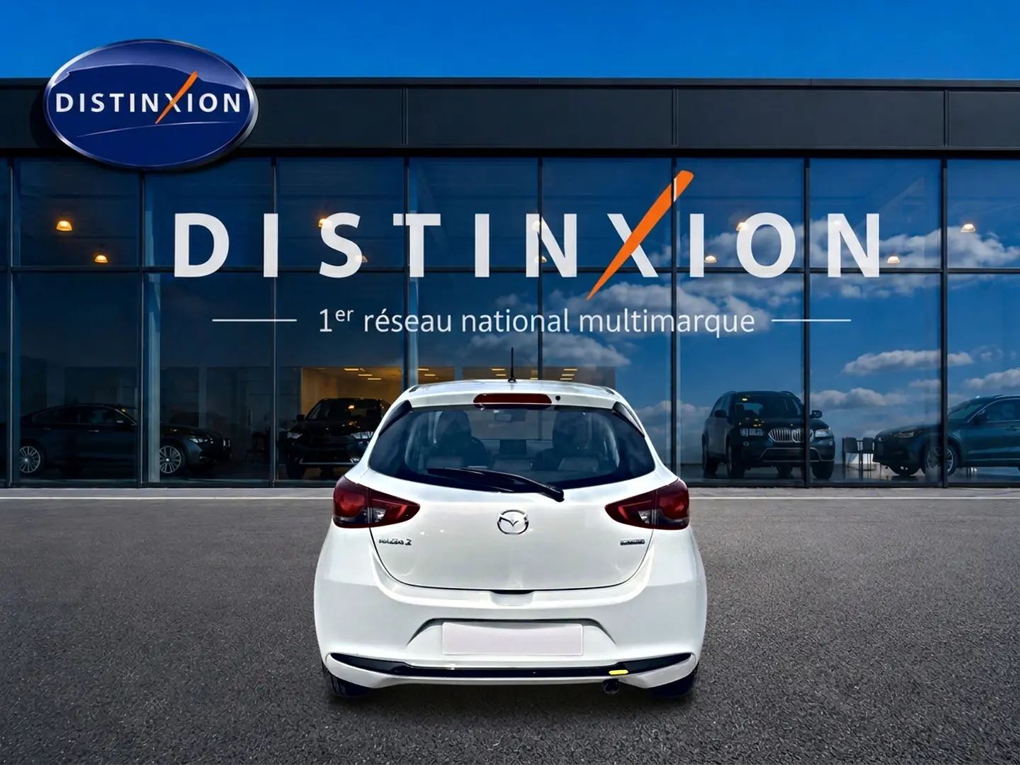 Vue arrière d'une Mazda2 blanche 2024 garée devant une vitrine avec logo Distinxion en arrière-plan.