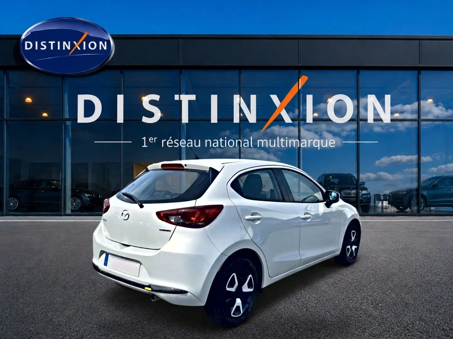 Vue 3/4 arrière droite d'une Mazda2 blanche 2024 garée devant une vitrine avec logo Distinxion.