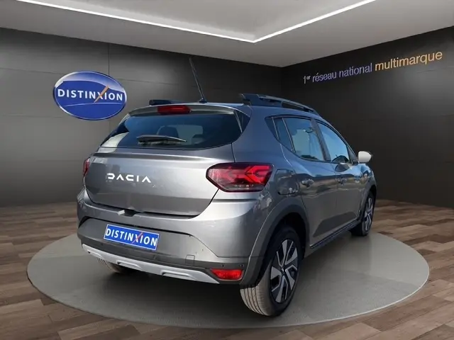 Vue 3/4 arrière droite d'une Dacia Sandero Stepway gris foncé avec rails de toit et feux arrière LED dans un showroom.