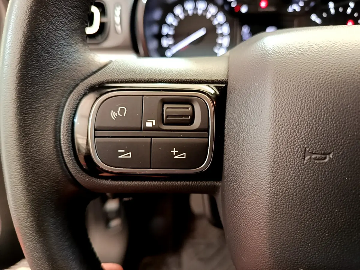 Gros plan sur les commandes gauche du volant cuir de la Citroën C3 gris foncé, avec tableau de bord flou en arrière-plan.