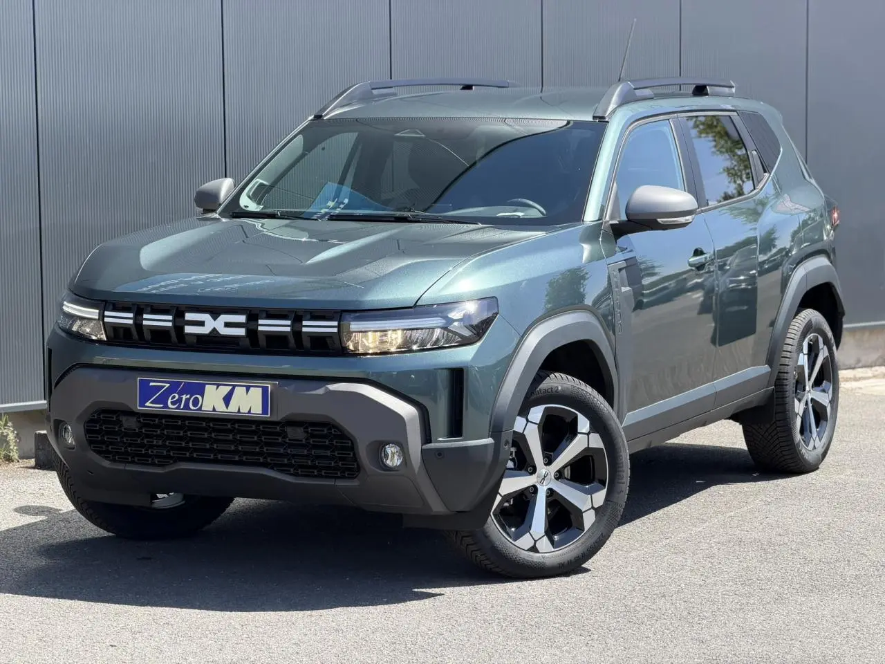 Vue 3/4 avant d'un Dacia Duster Hybrid-G 150 4x4 vert cèdre avec jantes alliage et barres de toit noires.