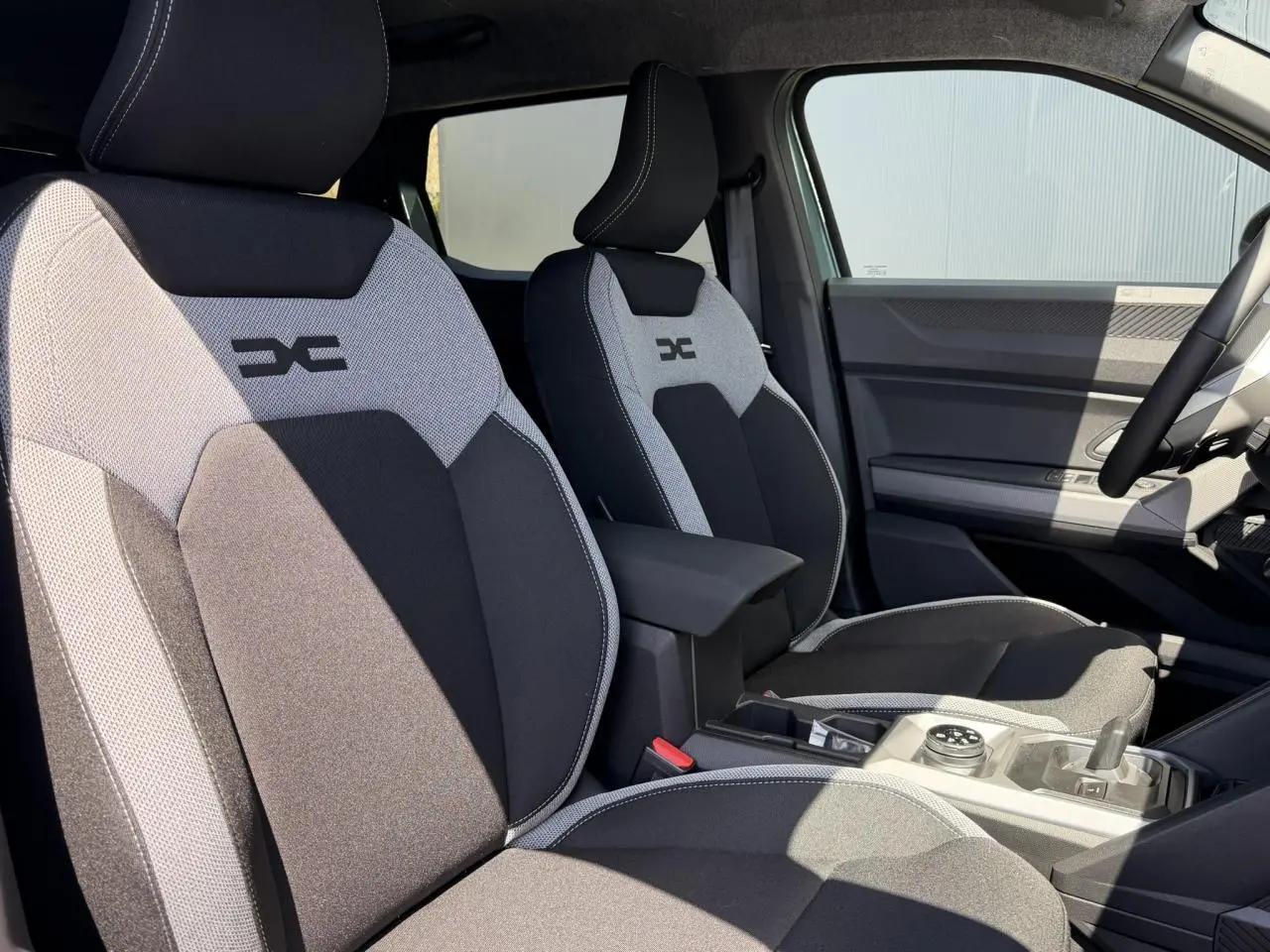 Vue intérieure des sièges avant gris et noir avec logo Dacia, montrant la console centrale et le volant côté droit.