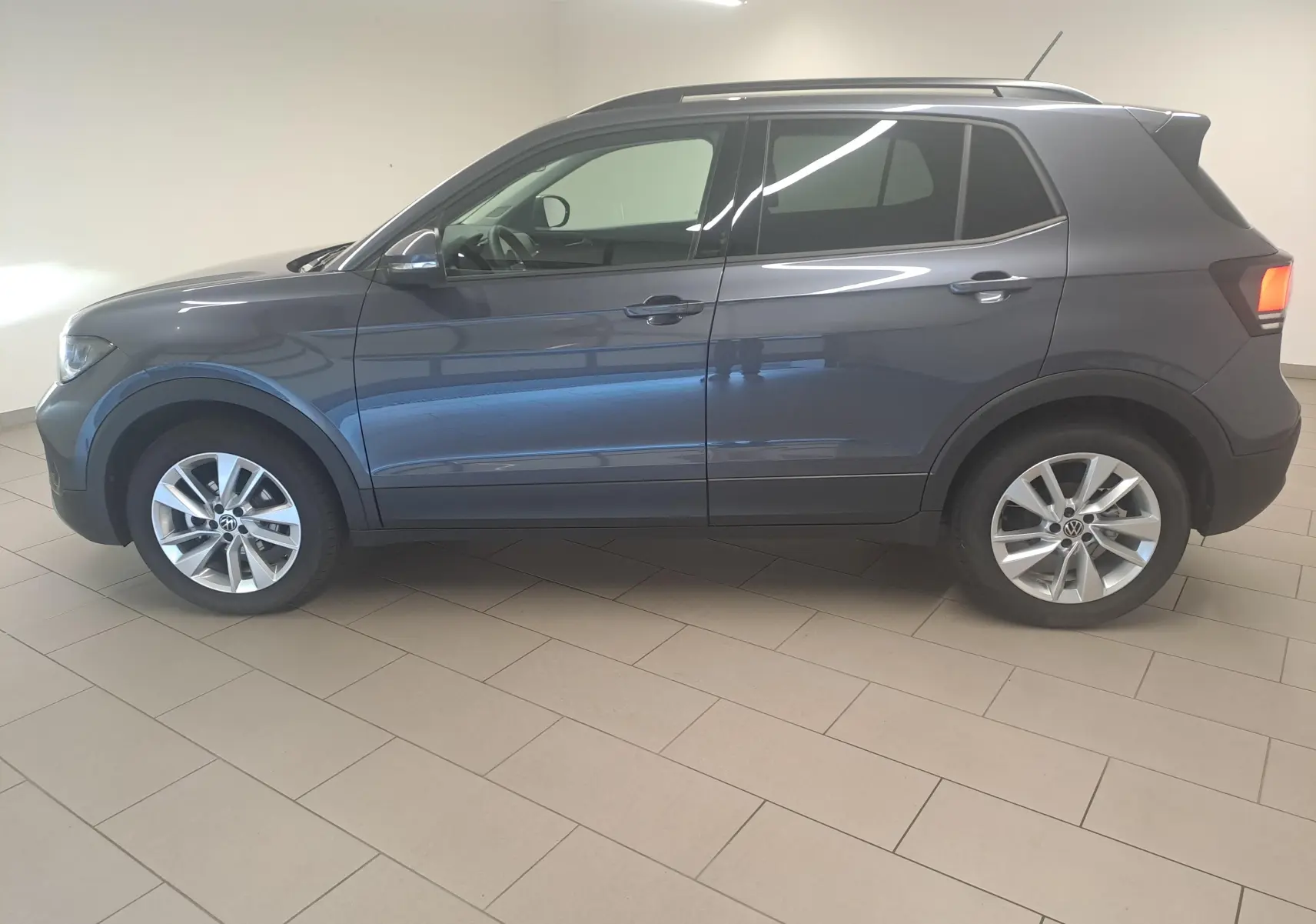Vue de profil côté gauche d'un Volkswagen T-Cross 2025 gris cendré métallisé avec jantes alliage et feux arrière allumés.