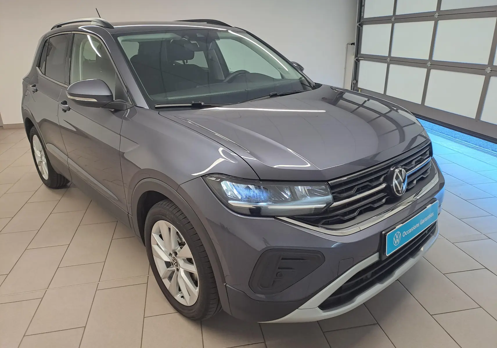 Volkswagen T-Cross gris cendré en 3/4 avant droit, avec calandre noire et feux LED allumés en intérieur showroom.