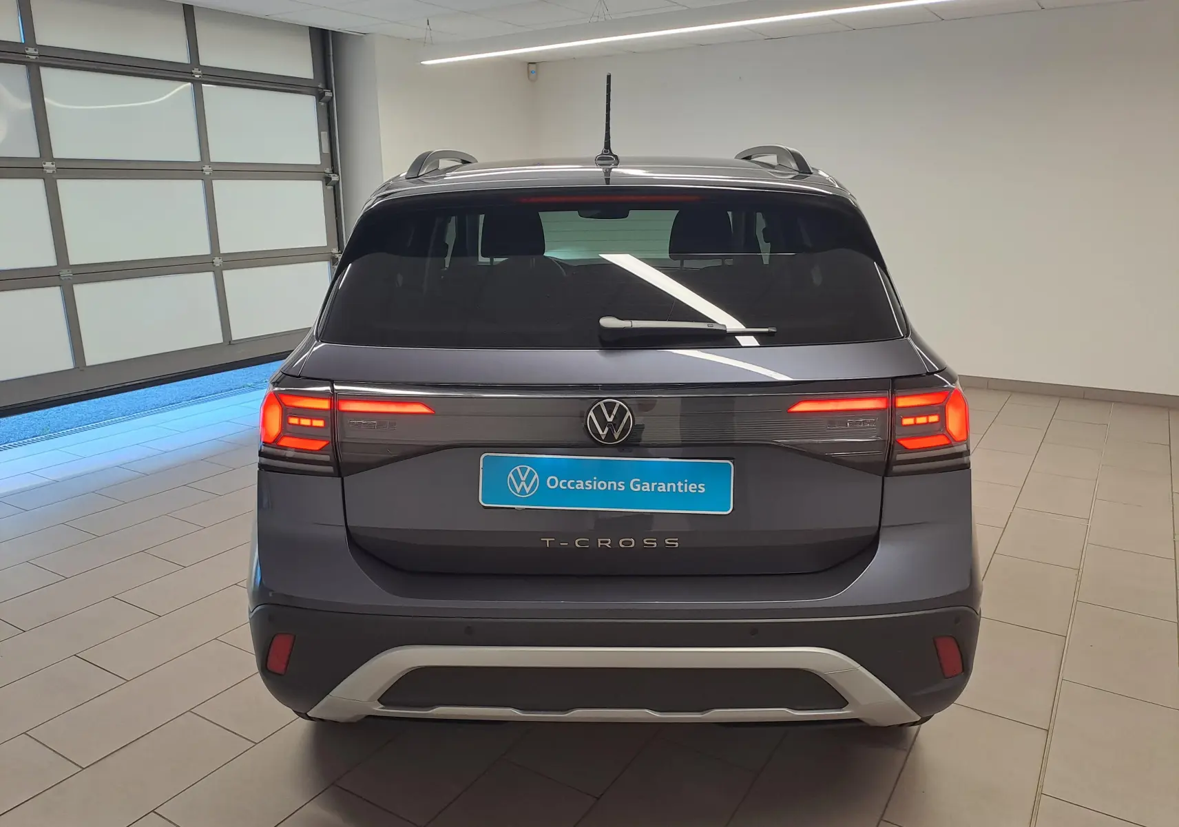 Vue arrière d'un Volkswagen T-Cross gris cendré 2025 avec feux allumés et plaque "Occasions Garantis" en intérieur showroom.